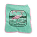 The Mote Marine Laboratory Raschel Blanket is a green cloth blanket with pink trim, showcasing an illustration of a spoonbill bird standing in water, surrounded by flying birds and trees in the background. The text “MOTE MARINE LABORATORY” appears below the image, reflecting a commitment to marine conservation.