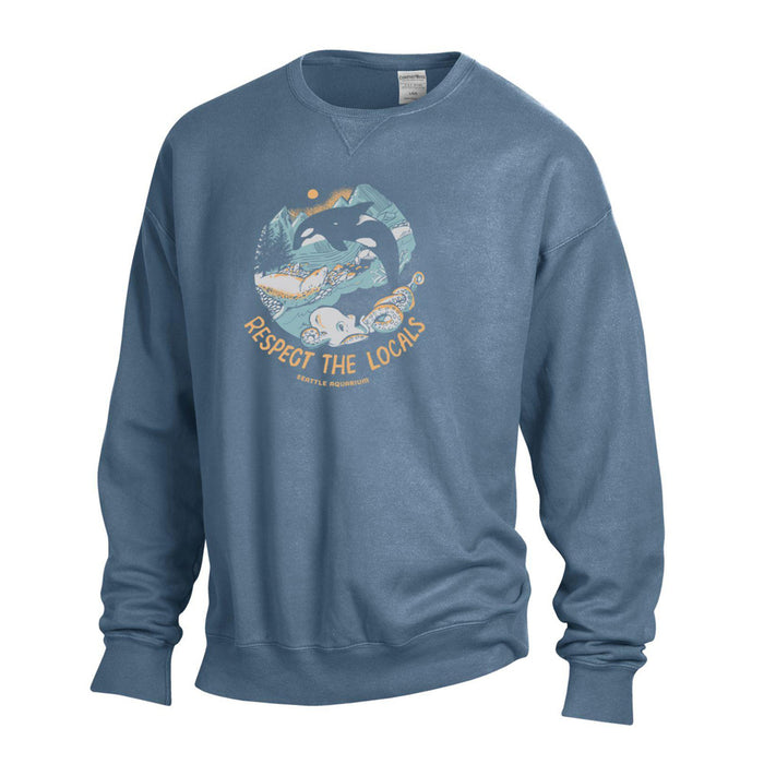 The Seattle Aquarium Respect the Locals Sweatshirt features a blue crewneck design with an illustration of an orca leaping over waves, framed by trees and mountains. Below the image is the text “RESPECT THE LOCALS” along with “Seattle, Washington.” This marine life sweatshirt makes a perfect ocean conservation gift for anyone who loves the Pacific Northwest.