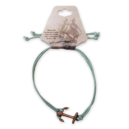 The Adjustable Cord Bracelet features a light blue cord and a small copper USS Constitution anchor charm, crafted from reclaimed copper. Displayed on a tag with a sailing ship, it celebrates the historic vessel’s legacy.