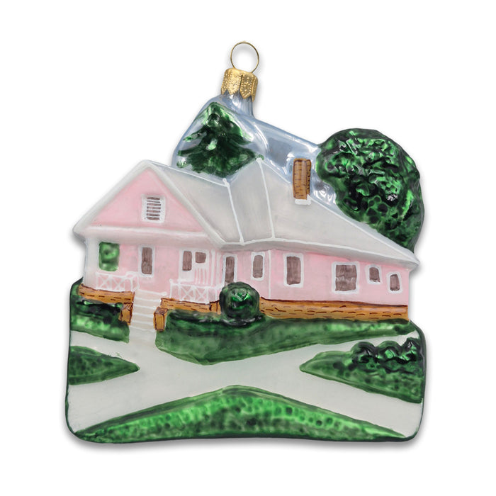 The Desert Botanical Garden Building Ornament [waiting] is a ceramic pink house with a white roof, green trees, and bushes. It includes a gold hanging loop, porch, and front paths—perfect for Southwest decor.