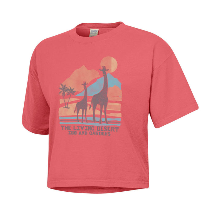 The Living Desert Zoo and Gardens Women’s Crop T-Shirt—a pink crop top featuring two giraffes, palm trees, mountains, and a sun with “The Living Desert Zoo and Gardens” text on the front—is perfect for stylishly supporting wildlife conservation.