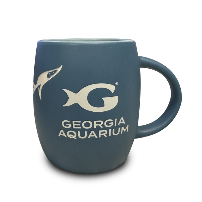 The Georgia Aquarium Whale Shark Etched Mug is a blue ceramic mug featuring an etched shark silhouette and the “G Georgia Aquarium” logo in white, with a large curved handle on the right.