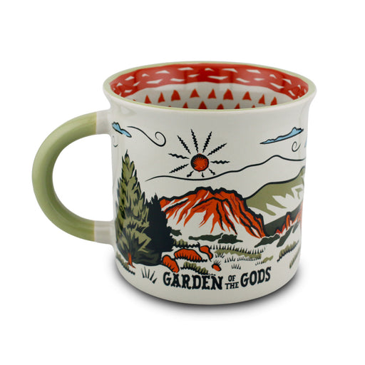 The Garden of the Gods Feeling Folksy Mug is a ceramic mug inspired by Colorado Springs, featuring a painted landscape of mountains, trees, and the sun. It has a green handle, a red patterned interior, and is labeled “Garden of the Gods.”.