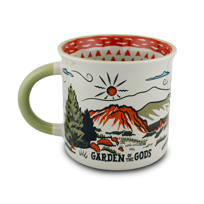 The Garden of the Gods Feeling Folksy Mug is a ceramic mug inspired by Colorado Springs, featuring a painted landscape of mountains, trees, and the sun. It has a green handle, a red patterned interior, and is labeled “Garden of the Gods.”.