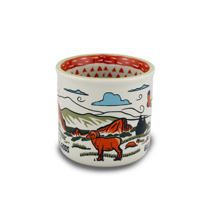 Introducing the Garden of the Gods Feeling Folksy Mini Mug—a ceramic cup adorned with stylized illustrations of mountains, a ram, trees, and a blue sky. Featuring earthy tones, bold outlines, and a red geometric pattern around the rim and interior, this mini mug makes for an ideal Colorado souvenir or keepsake from Garden of the Gods.