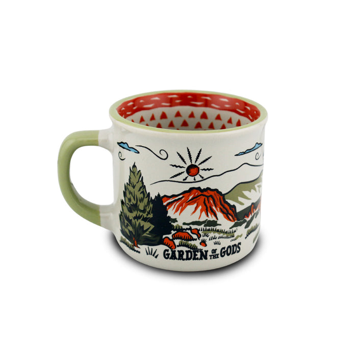 The Garden of the Gods Feeling Folksy Mini Mug is a ceramic mug with a green handle, showcasing a colorful landscape illustration of mountains, trees, and a red sun along with the “Garden of the Gods” text. Its red interior features a white triangular pattern, making it an ideal Colorado souvenir or mini espresso mug for your collection.