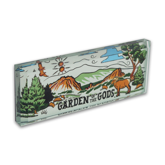 The Garden of the Gods Feeling Folksy Glass Magnet is a rectangular glass souvenir featuring an illustrated landscape with mountains, trees, a ram, the sun, clouds, vibrant red rock formations, and “Garden of the Gods” text.