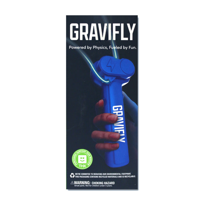 A hand holds a blue Genius at Play GraviFly Rope Launcher with gravity-defying design. The packaging says: GraviFly. Powered by Physics, Fueled by Fun. Icons show eco commitment and a choking hazard warning appears at the bottom.