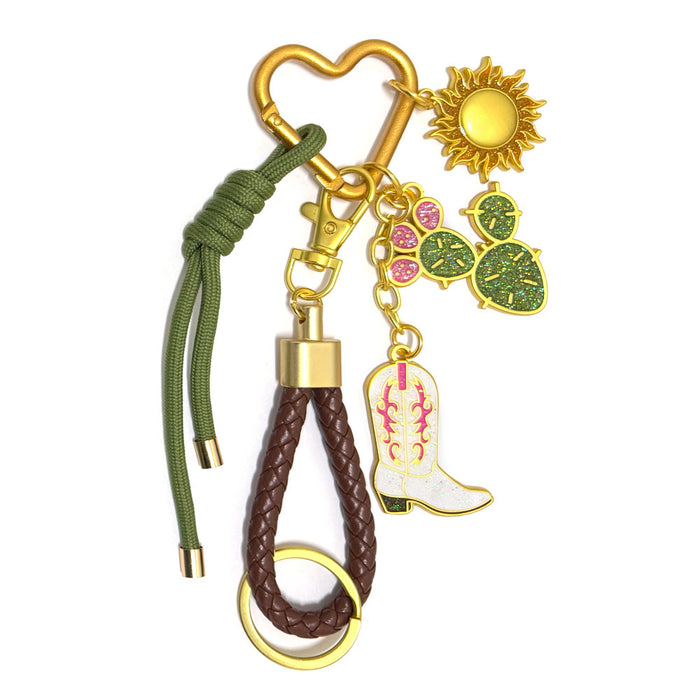The Desert Carabiner Bag Clip features a gold keychain with a heart clip, sun and cactus charms, pink paw print, white cowboy boot, braided brown leather loop, and green cord—a stylish accessory for your handbag.