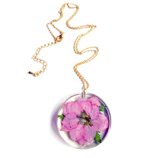 The Sakura Cherry Petal Necklace is a gold chain with a round, clear pendant that showcases a preserved pink flower and green leaves, inspired by Japanese jewelry for a timeless, nature-inspired look.