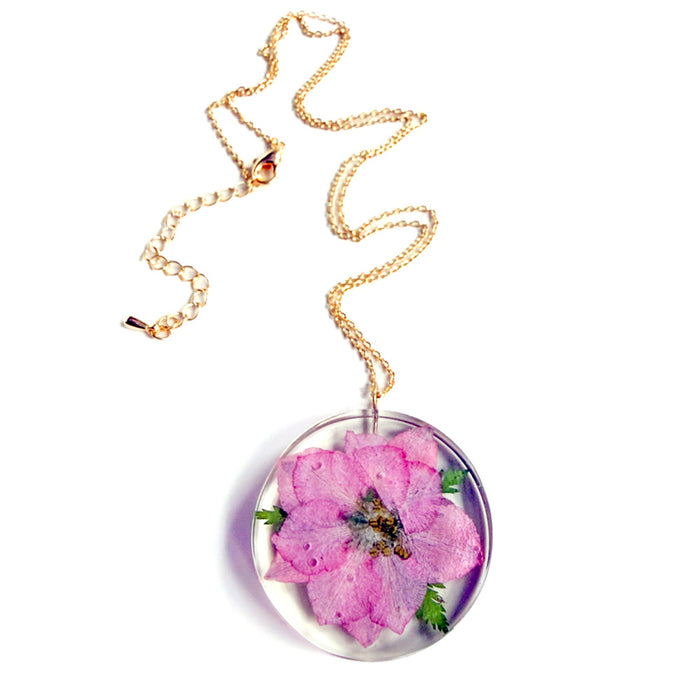 The Sakura Cherry Petal Necklace is a gold chain with a round, clear pendant that showcases a preserved pink flower and green leaves, inspired by Japanese jewelry for a timeless, nature-inspired look.