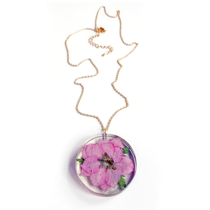 The Sakura Cherry Petal Necklace is a gold chain with a round pendant, featuring a pressed pink flower encased in clear resin for a delicate and timeless Japanese-inspired look.