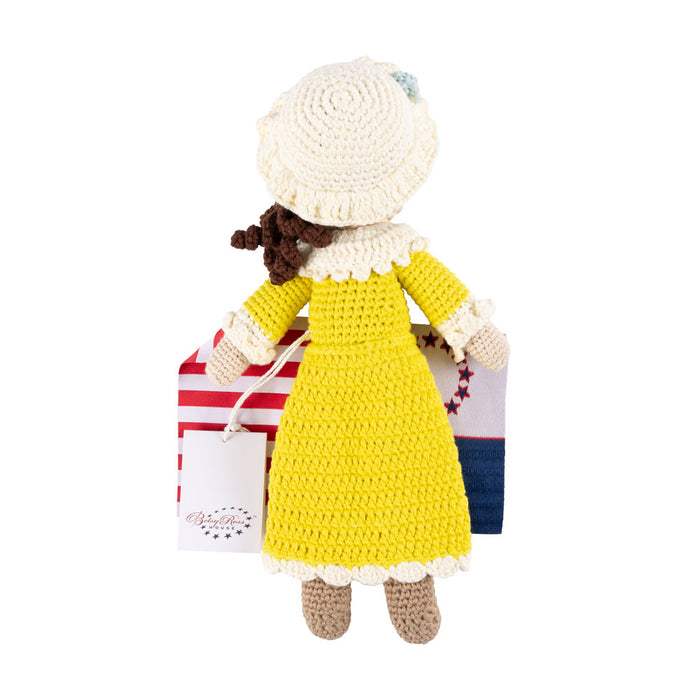 The Betsy Ross House Crocheted Doll, featuring a yellow dress and cream bonnet, is a handmade keepsake displayed in front of a folded Betsy Ross-style American flag.
