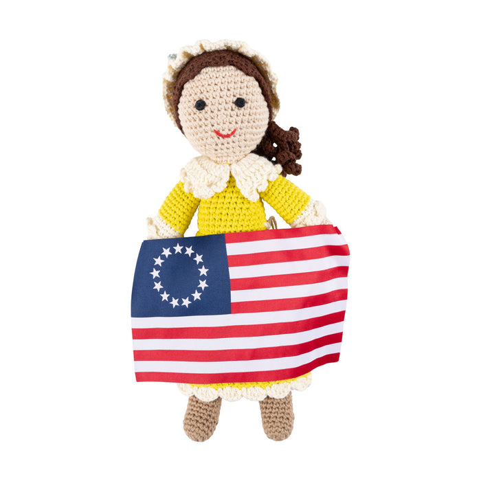 The Betsy Ross House Crocheted Doll is a charming handmade keepsake, featuring a doll in a yellow dress and white bonnet holding a Betsy Ross American flag with 13 stars in a circle and 13 red and white stripes—a perfect addition to any collection.