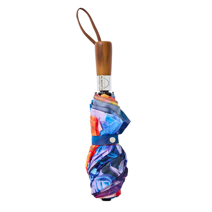 Closed Painter’s Palette Travel Umbrella featuring a multicolor abstract pattern, a wooden handle, and a leather wrist strap.