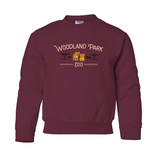 A maroon Woodland Park Zoo Baby Lion Crew Youth Sweatshirt featuring two lions, tree silhouettes, and a wildlife conservation theme printed on the front.