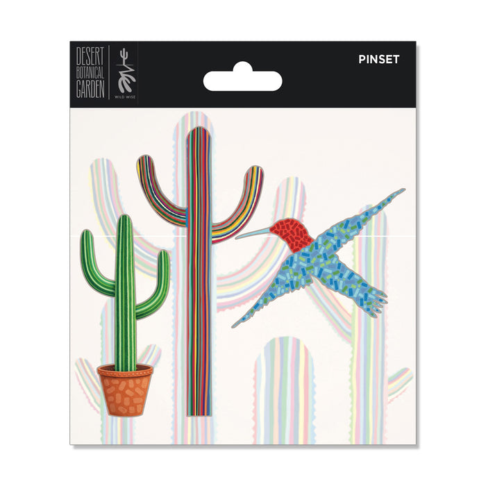 The Desert Botanical Garden Will Beger Pin Set features desert flora designs—a potted cactus, a tall rainbow-striped cactus, and a blue-and-red hummingbird—displayed on a white background with shadow outlines reminiscent of the Desert Botanical Garden.