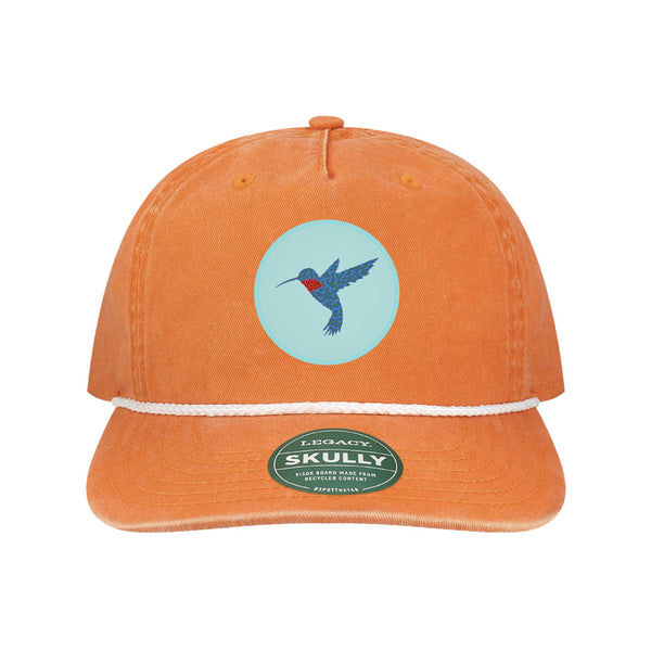 The Desert Botanical Garden Will Beger Hummingbird Patch Cap is an orange cap featuring a white rope above the brim and a circular patch with a blue and red hummingbird, designed by Will Beger. It also includes a green Skully brand sticker on the brim.