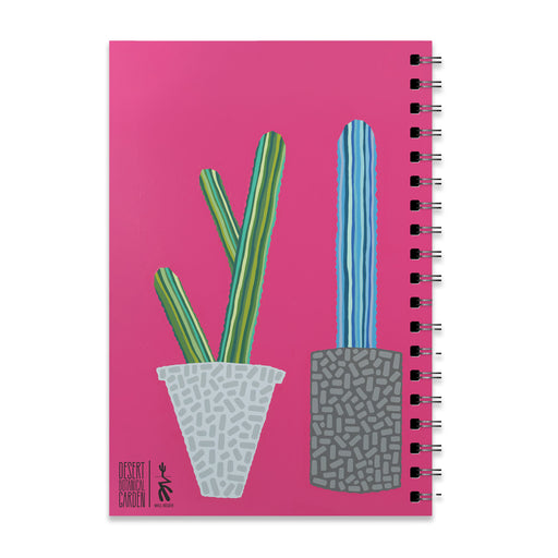 The Desert Botanical Garden Will Beger Green & Blue Cactus Notebook is a spiral-bound notebook with a vibrant pink cover, decorated with illustrations inspired by the Desert Botanical Garden. The design features two cacti in patterned pots: a green cactus in a white pot on the left, and a taller blue and green cactus in a gray pot on the right, evoking Will Beger’s playful artistic style.