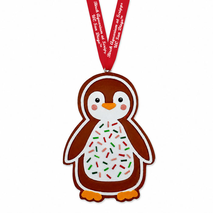 Penguin-shaped cookie ornament decorated with white icing and red and green sprinkles, hanging from a red ribbon labeled Birch Aquarium at Scripps UC San Diego.
