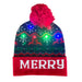 Navy blue Holiday Lights Beanie with a red cuff reading MERRY, patterned with snowflakes and reindeer, topped with a red pom-pom and glowing multicolored LED lights.