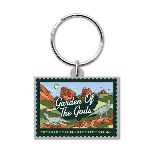 Garden of the Gods Colorado 150th Keychain featuring a rectangular design with an illustrated stamp motif that depicts red rock formations, dinosaurs, and lush greenery—a tribute to the iconic Colorado landmark. The keychain displays the text “Garden of the Gods” and “Sesquisesquiquincentennial,” marking a special 150th anniversary celebration. A silver keyring is attached at the top.