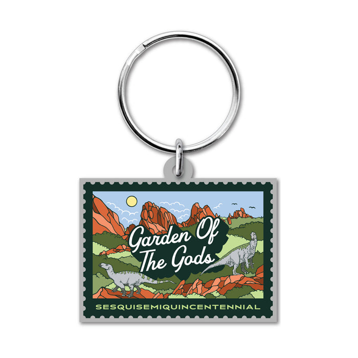 Garden of the Gods Colorado 150th Keychain featuring a rectangular design with an illustrated stamp motif that depicts red rock formations, dinosaurs, and lush greenery—a tribute to the iconic Colorado landmark. The keychain displays the text “Garden of the Gods” and “Sesquisesquiquincentennial,” marking a special 150th anniversary celebration. A silver keyring is attached at the top.