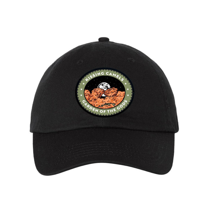 The Garden of the Gods Kissing Camels Cap is a black hat adorned with a patch depicting brown rock formations, a moon, stars, and the words “KISSING CAMELS GARDEN OF THE GODS,” evoking the rugged beauty of Colorado’s Garden of the Gods.