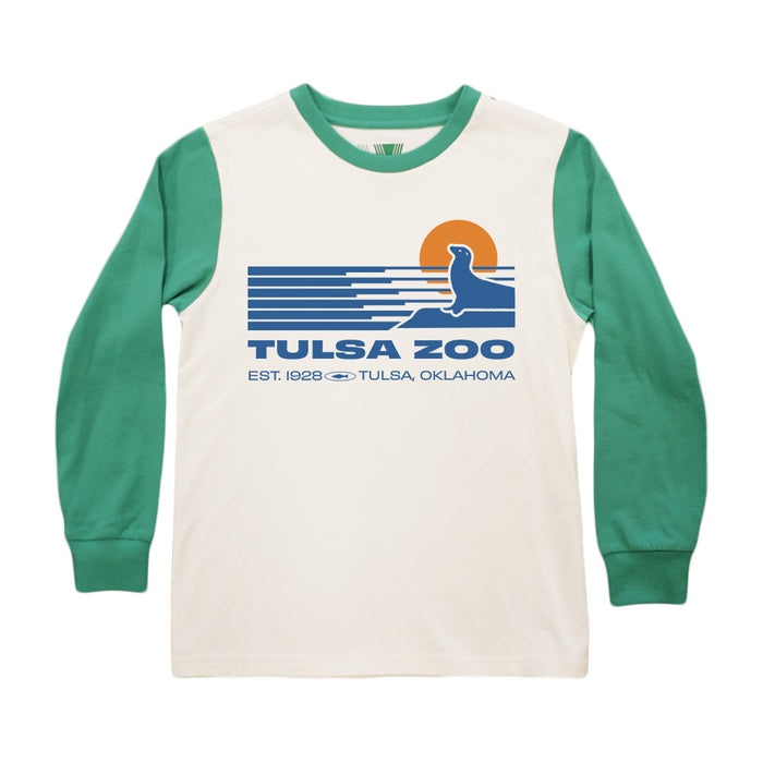 The Tulsa Zoo Sea Lion Sporty Pop Youth T-Shirt features green long sleeves, a white organic cotton body, and a front graphic of a seal, setting sun, blue waves, and “Tulsa Zoo Est. 1928 Tulsa, Oklahoma.”.