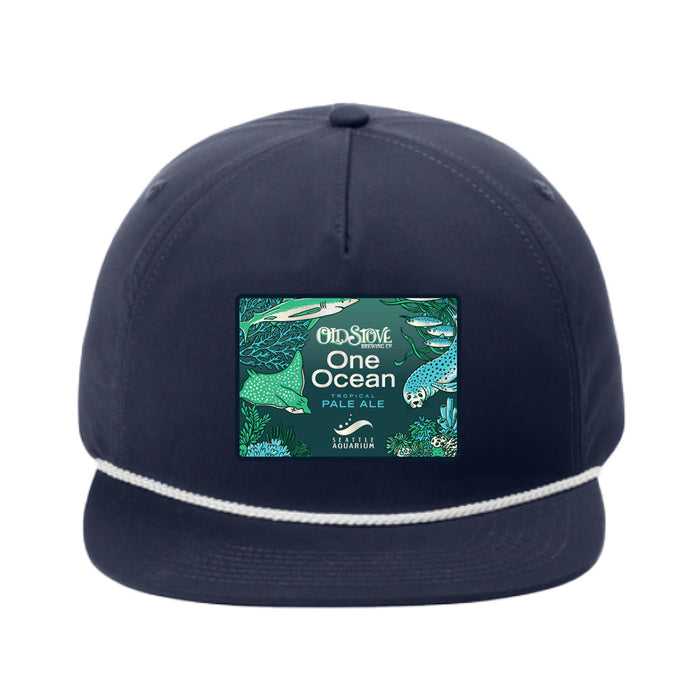 The Seattle Aquarium Old Stove Brewing Co. Rope Cap is a navy blue cap with a white rope detail above the brim, showcasing a vibrant patch that reads “One Ocean Pale Ale” alongside ocean-themed illustrations. This cap was created in collaboration with Old Stove Brewing Co., inspired by the spirit of the Seattle Aquarium.
