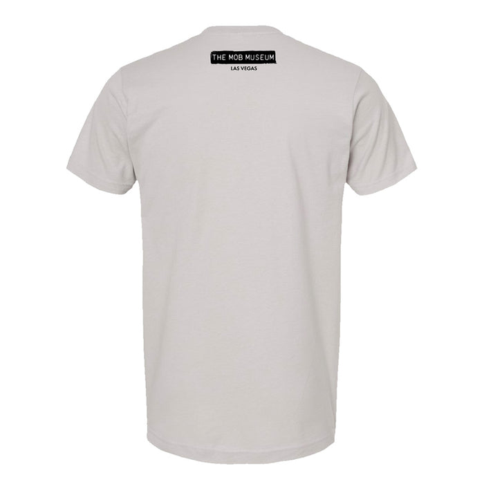 Back view of a plain light gray The Mob Museum Happy Face T-Shirt featuring a small black rectangular label near the neck that reads THE MOB MUSEUM LAS VEGAS in white, uppercase letters—a perfect Las Vegas souvenir for fans of history and intrigue.