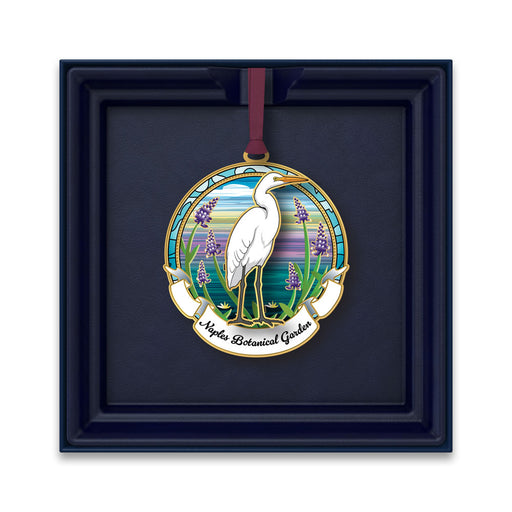 The Naples Botanical Garden Egret Ornament showcases a white heron standing amid purple flowers beneath a vibrant sky, with “Naples Botanical Garden” inscribed below—an ideal Florida keepsake presented in a dark blue box.