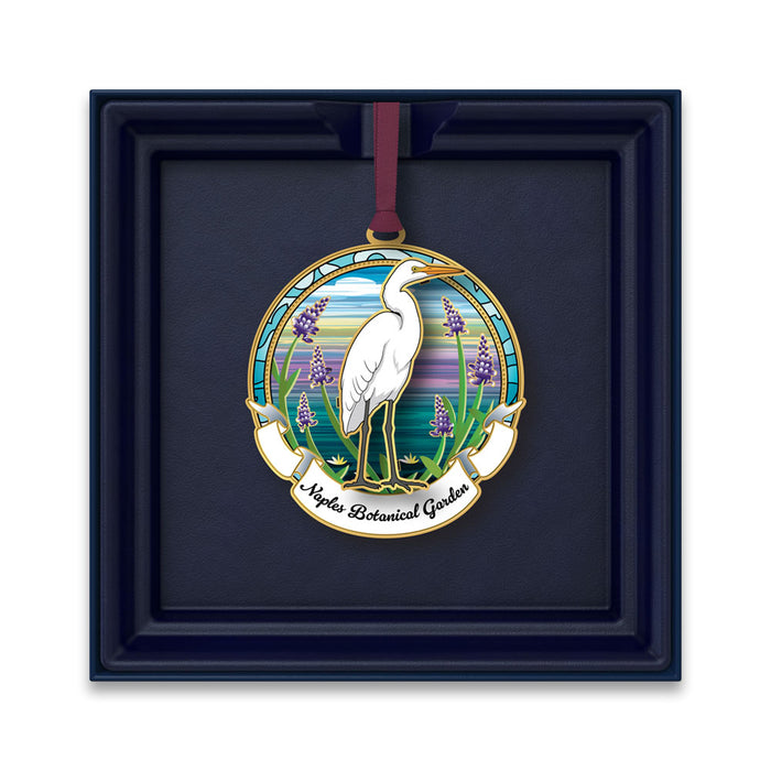 The Naples Botanical Garden Egret Ornament showcases a white heron standing amid purple flowers beneath a vibrant sky, with “Naples Botanical Garden” inscribed below—an ideal Florida keepsake presented in a dark blue box.