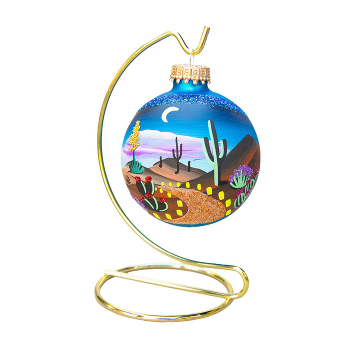 Desert Botanical Garden Brenda J Luminaria 2026 Ornament featuring a blue glass hand-painted desert landscape with saguaro cacti and a path, hanging on a gold metal stand.