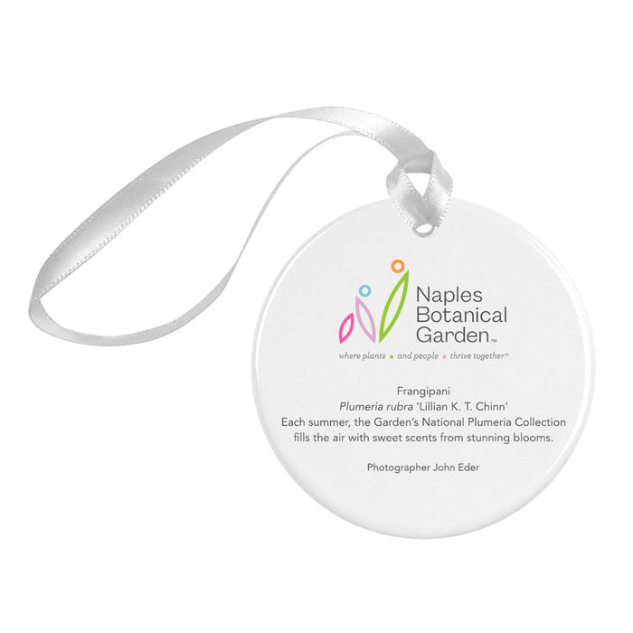 The Naples Botanical Garden John Eder Ornament is a round white porcelain ornament accented with a silver ribbon. It features the Naples Botanical Garden logo and text about the Frangipani flower, along with a photography credit to John Eder. This ornament beautifully captures the essence of Naples Botanical Garden.