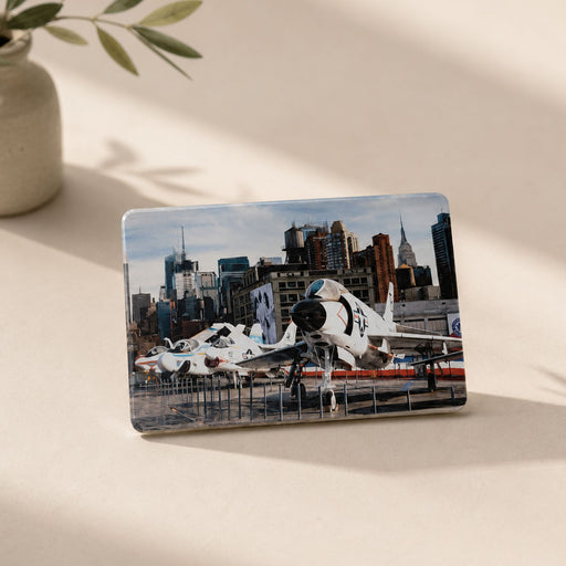 A rectangular Intrepid Museum Planes Photo Magnet features fighter jets displayed outdoors with a city skyline in the background, capturing the excitement of the museum for aviation enthusiasts. The magnet is propped up on a light surface next to a vase with green leaves.