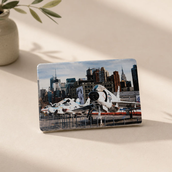 A rectangular Intrepid Museum Planes Photo Magnet features fighter jets displayed outdoors with a city skyline in the background, capturing the excitement of the museum for aviation enthusiasts. The magnet is propped up on a light surface next to a vase with green leaves.