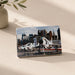 A rectangular Intrepid Museum Planes Photo Magnet features fighter jets displayed outdoors with a city skyline in the background, capturing the excitement of the museum for aviation enthusiasts. The magnet is propped up on a light surface next to a vase with green leaves.