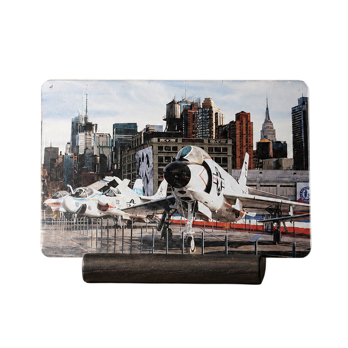 A variety of jet aircraft are showcased outdoors on a platform at the Intrepid Museum, set against the backdrop of towering city skyscrapers and a clear sky—an ideal view for aviation enthusiasts or as featured on the Intrepid Museum Planes Photo Magnet.
