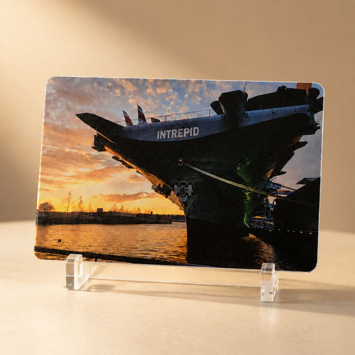 A printed photo of the USS Intrepid aircraft carrier at sunset stands upright on a clear plastic display holder, making it a perfect companion piece to the Intrepid Museum Ship Photo Magnet or a memento from the Intrepid Sea, Air & Space Museum.