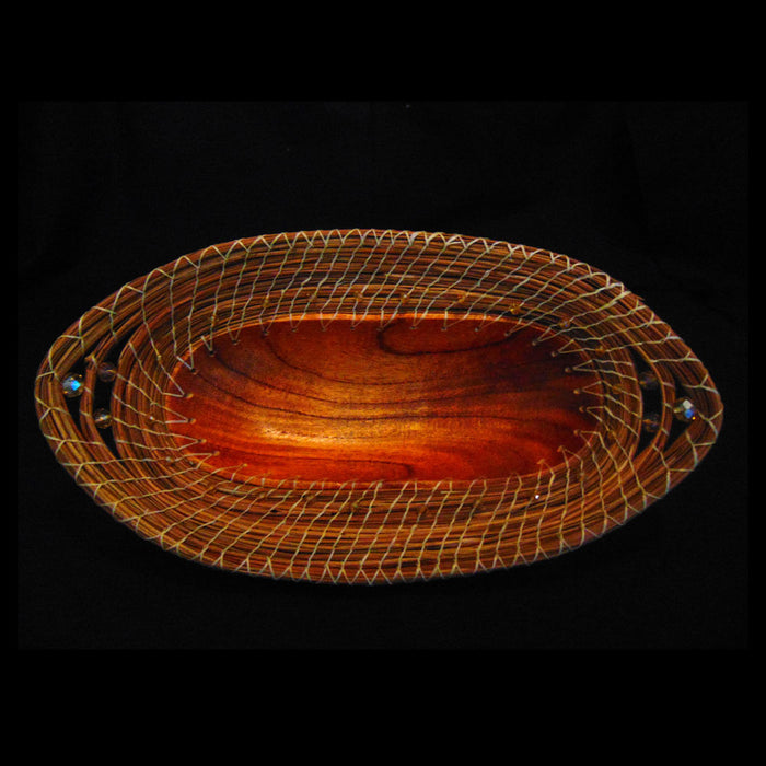 Introducing the Large Pine Needle Oval Basket: a handcrafted piece featuring an elegant wooden oval bowl highlighted by intricate, light-colored wirework along its rim and finished with two turquoise beads set on opposite ends. This striking, sustainable home decor item stands out beautifully against a black background.