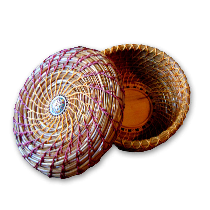 The Multicolor Pine Needle Basket is a round, handcrafted basket with a fitted lid, featuring eco-friendly natural and reddish-brown fibers woven in a circular pattern and an intricate design at the center of the lid.