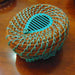 The Turquoise Pine Needle Basket is a small, round, handwoven piece crafted with brown fibers and accented by blue threading. This eco-friendly decor item features a lid adorned with a slotted black and blue oval design at its center.