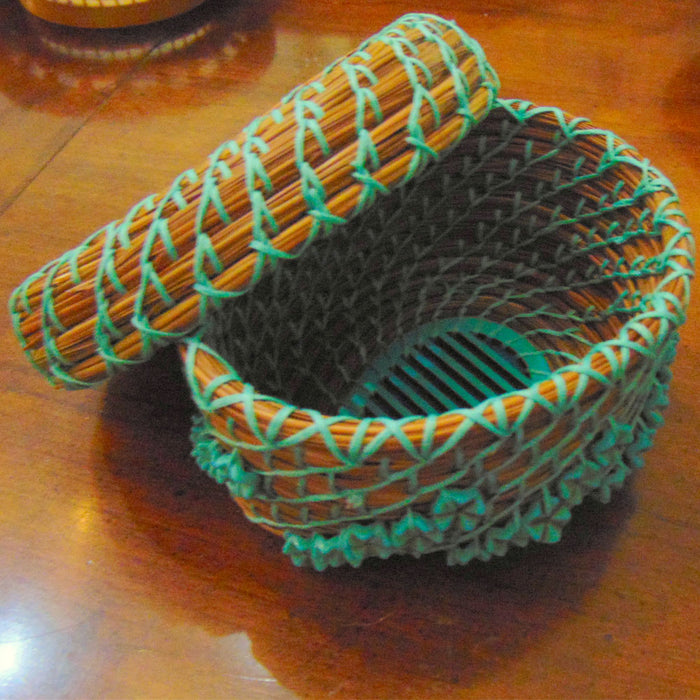 A small, round Turquoise Pine Needle Basket with a matching lid, handwoven from brown and teal materials, sits open on a wooden surface—an eco-friendly decor piece.