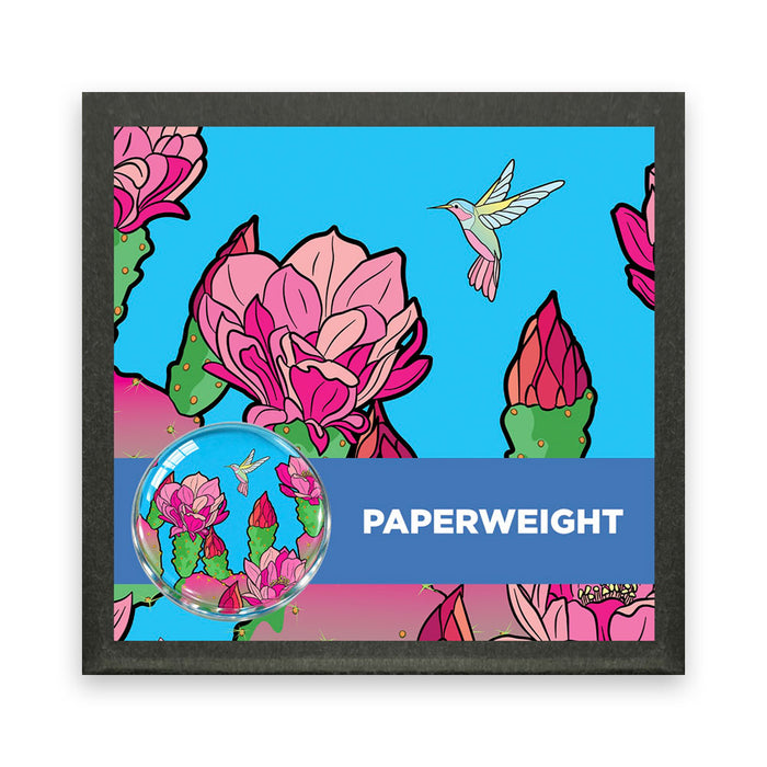 Desert Botanical Garden Superbloom Paper Weight, a round glass piece featuring a hummingbird and pink cactus flowers on a blue background, displayed against a matching illustrated card.