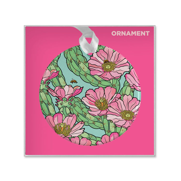 Desert Botanical Garden Superbloom Ornament, a round porcelain piece featuring a light blue illustration of pink cactus flowers and green cacti, hanging by a white ribbon against a bright pink background.