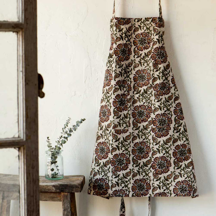 Elysian Block Print Apron in chambray cotton with an intricate floral pattern hanging against a white wall next to a wooden stool holding a small glass jar with a sprig of greenery.