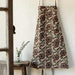 Elysian Block Print Apron in chambray cotton with an intricate floral pattern hanging against a white wall next to a wooden stool holding a small glass jar with a sprig of greenery.