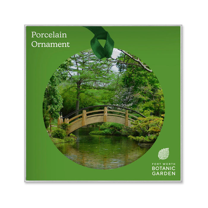 Square packaging for a Fort Worth Botanical Garden Ornament, featuring a circular image of a wooden arched bridge over a pond in the garden.