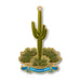 A gold-toned Desert Botanical Garden Saguaro Ornament shaped like a cactus in a desert landscape, featuring a blue ribbon at the bottom that reads Desert Botanical Garden.